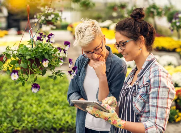 Two women in a garden center, engrossed in a tablet, discussing plants and gardening tips.