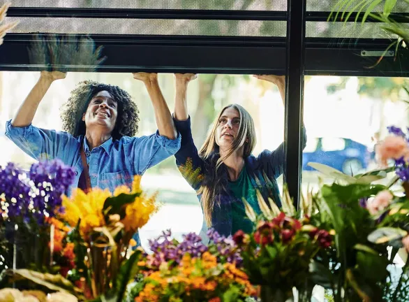 Two women gazing out of a window at colorful flowers in bloom.